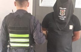 Oficial de policía con chaleco táctico sostiene a un detenido con capuchón negro en una comisaría.