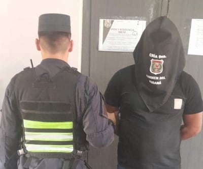 Oficial de policía con chaleco táctico sostiene a un detenido con capuchón negro en una comisaría.