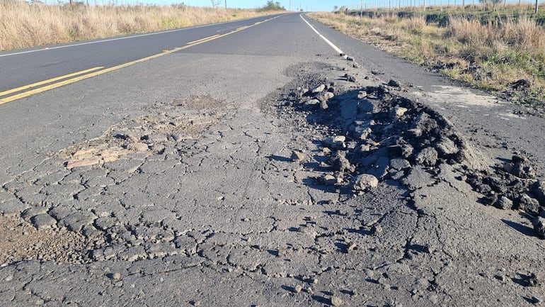 Carretera en mal estado con grietas y bultos, rodeada de hierbas secas y vegetación escasa.