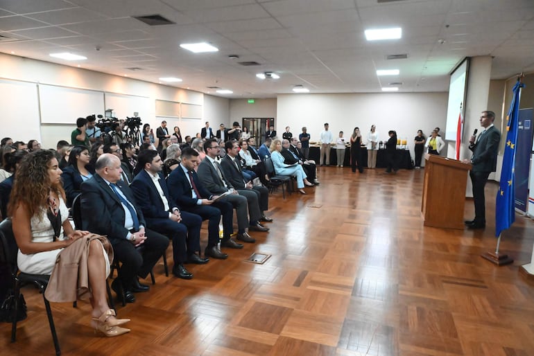 Ambiente formal con Marco Riquelme y Katja Afheldt en trajes formales, escuchando a un orador en un podio, rodeados de audiencia.