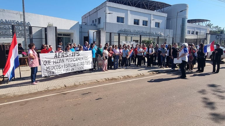 Protesta pacífica con aproximadamente 40 personas sosteniendo carteles sobre salud frente al IPS, bajo un día soleado.