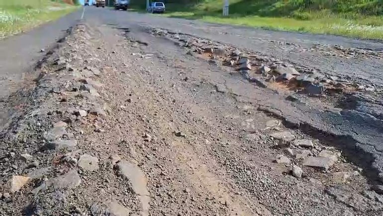Carretera deteriorada con escombros, sin personas visibles; vehículos circulando al fondo.