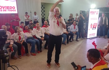 Arnoldo Wiens en el centro con micrófono, rodeado de personas en camisas blancas y pañuelos rojos, en un ambiente formal.