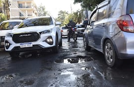 Motociclista de pie con chaqueta negra y casco, frente a vehículos detenidos en calle con baches y charcos de agua.