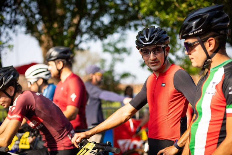 Ciclistas conversan, uno con camiseta roja y el otro en colores verde, blanco y rojo, rodeados de árboles y bicicletas numeradas.