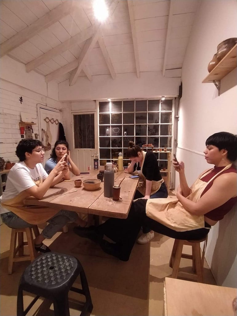 Cuatro personas sentadas en mesa de madera. Dos conversan, mientras otras dos miran sus teléfonos. Taller con luz natural.