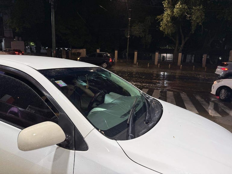 Automóvil blanco dañado con parabrisas fracturado, mojadura por lluvia y otros vehículos en ambiente urbano.