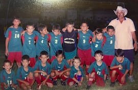 Niños con camisetas verdes y rojas sonríen junto a Orlando Gill y Lucho Mendoza en un campo de fútbol al aire libre.