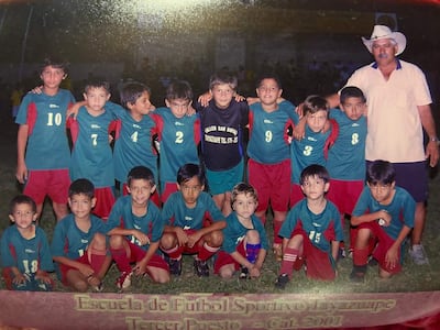 Niños con camisetas verdes y rojas sonríen junto a Orlando Gill y Lucho Mendoza en un campo de fútbol al aire libre.