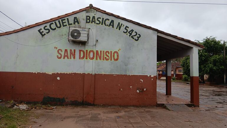 Fachada de la Escuela Básica N° 5423 San Dionisio, con nombre visible en rojo, y suelo mojado tras la lluvia.