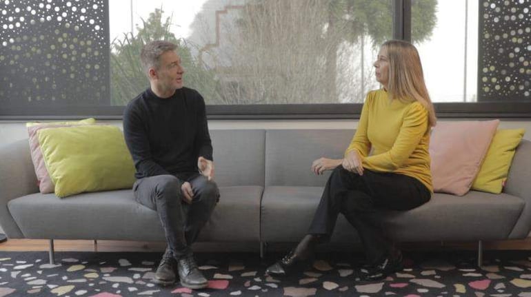 Dos adultos sentados en un sofá, uno con suéter negro y pantalones grises, el otro con blusa amarilla, conversando en un ambiente acogedor.