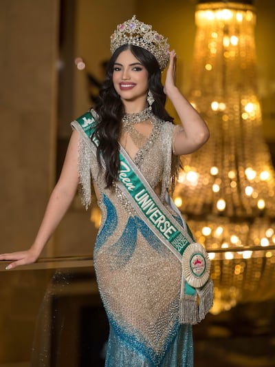 Katya Jel sonríe con vestido plateado y corona en un ambiente festivo iluminado por una lámpara de araña.