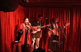 Vocalista mujer canta al micrófono, rodeada de dos hombres con guitarras, en un ambiente cálido y acogedor.