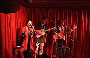 Vocalista mujer canta al micrófono, rodeada de dos hombres con guitarras, en un ambiente cálido y acogedor.