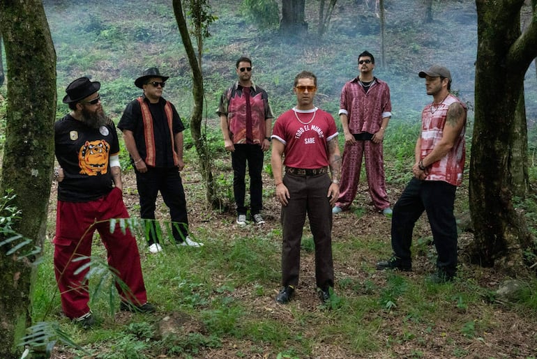 Seis hombres en un entorno natural se agrupan, visten ropas coloridas, uno destaca con una camiseta que dice 'TODO EL MUNDO SABE'.