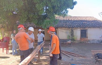 Hombres con cascos naranjas y gris, conversan y organizan materiales en la Casa de la Cultura, bajo un cielo despejado.