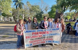 Grupo de bioquímicos con batas blancas y chaquetas sostienen un cartel en un parque, expresando reclamos laborales.