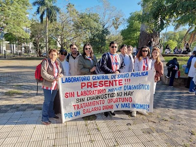 Grupo de bioquímicos con batas blancas y chaquetas sostienen un cartel en un parque, expresando reclamos laborales.