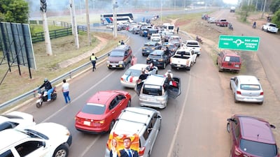 Multitud en carretera con banner político, personas de pie sobre vehículos, una persona con camiseta a rayas y ambiente congestionado.