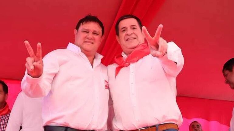 Hernán Rivas y Horacio Cartes en camisas blancas, levantan dos dedos en señal de paz, en un ambiente festivo de color rojo.