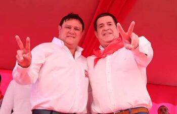 Hernán Rivas y Horacio Cartes en camisas blancas, levantan dos dedos en señal de paz, en un ambiente festivo de color rojo.