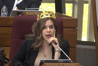 Mujer con corona dorada sentada, hablando en sillón de recinto legislativo, gesticulando con la mano cerca de la boca.