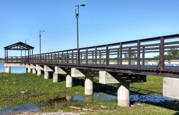 Muelle de madera elevado sobre el agua, rodeado de vegetación verde y farolas, en un día soleado.