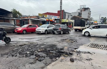 Motocicleta detenida en calle deteriorada, con vehículos en movimiento; ambiente nublado y comercial visible en fondo.