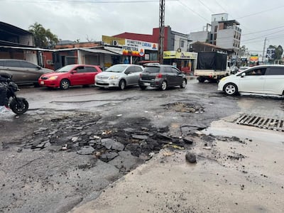 Motocicleta detenida en calle deteriorada, con vehículos en movimiento; ambiente nublado y comercial visible en fondo.