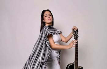 Mujer de pie con camiseta blanca y pantalones claros, sosteniendo guitarra con expresión confiada en ambiente de estudio.