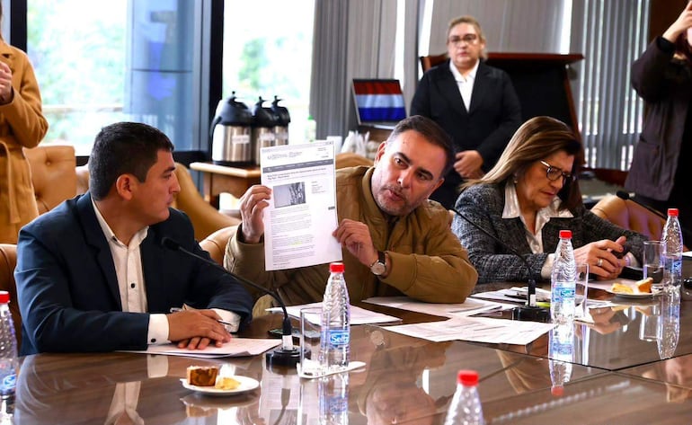 Claudia Centurión observa desde su asiento, mientras Ever Villalba sostiene un documento y explica al grupo en un ambiente laboral.