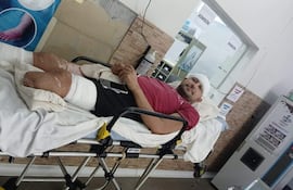 Hombre en camilla con venda en la cabeza y lesiones visibles, usando un teléfono móvil en un ambiente médico.