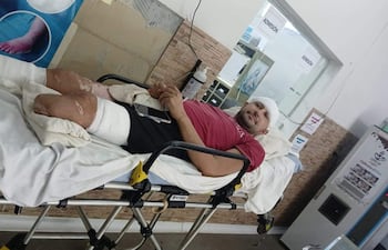 Hombre en camilla con venda en la cabeza y lesiones visibles, usando un teléfono móvil en un ambiente médico.