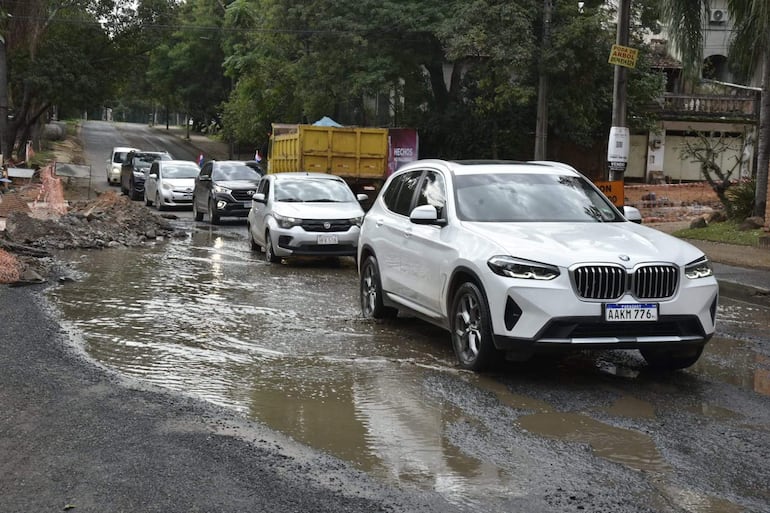 Varios vehículos en fila sobre un pavimento dañado y con agua, rodeados de árboles y edificaciones.