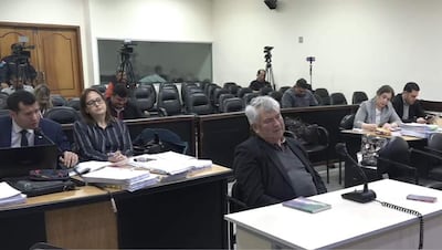Hombre mayor con cabello canoso reflexiona en un tribunal, acompañado de un abogado en traje que escribe en una laptop.