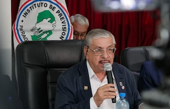 Isaías Fretes, en chaqueta oscura, habla con micrófono en mano, rodeado de un ambiente formal y una pared roja con el logo del IPS.
