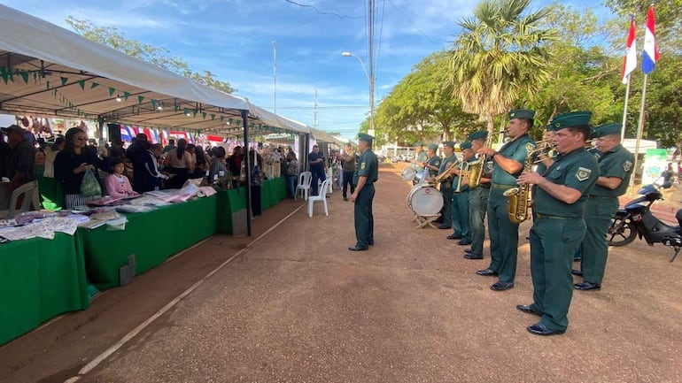 Músicos en uniformes verdes tocan instrumentos de viento en una feria, rodeados de stands y un ambiente festivo.