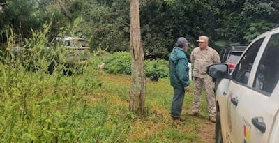 Hombres conversan en la naturaleza; uno con camisa camuflada y otro en abrigo verde, rodeados de vehículos todoterreno.