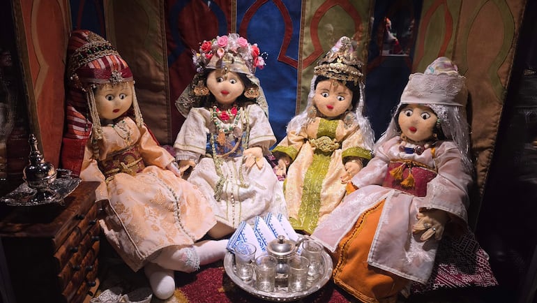 Cuatro muñecas con trajes tradicionales en círculo, acompañadas de un juego de té en un ambiente festivo colorido.