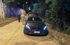 Dos suboficiales uniformados junto a un coche Mercedes-Benz oscuro, revisando la parte trasera del vehículo en un ambiente nocturno.
