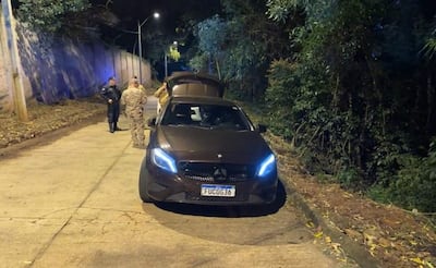 Dos suboficiales uniformados junto a un coche Mercedes-Benz oscuro, revisando la parte trasera del vehículo en un ambiente nocturno.