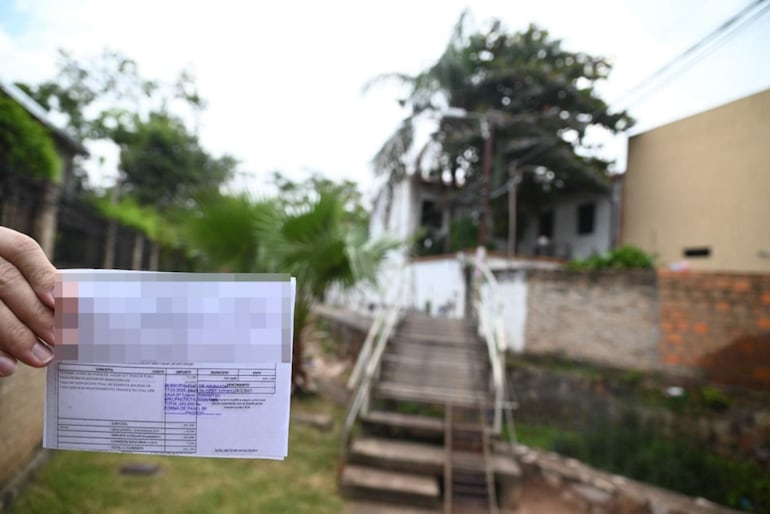 Un hombre sostiene un recibo en primer plano, con una casa sencilla y vegetación de fondo en un área residencial.