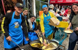 Cinco cocineros en delantales azules, uno con gorra negra y otro con gorro azul, fríen alimentos en un evento comunitario al aire libre.