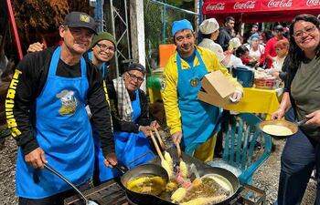 Cinco cocineros en delantales azules, uno con gorra negra y otro con gorro azul, fríen alimentos en un evento comunitario al aire libre.