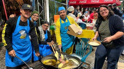 Cinco cocineros en delantales azules, uno con gorra negra y otro con gorro azul, fríen alimentos en un evento comunitario al aire libre.