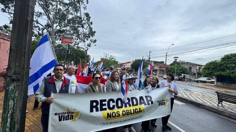 Grupo de adultos con banderas paraguaya e israelí, algunos con sombreros, avanzan en marcha por una calle nublada.