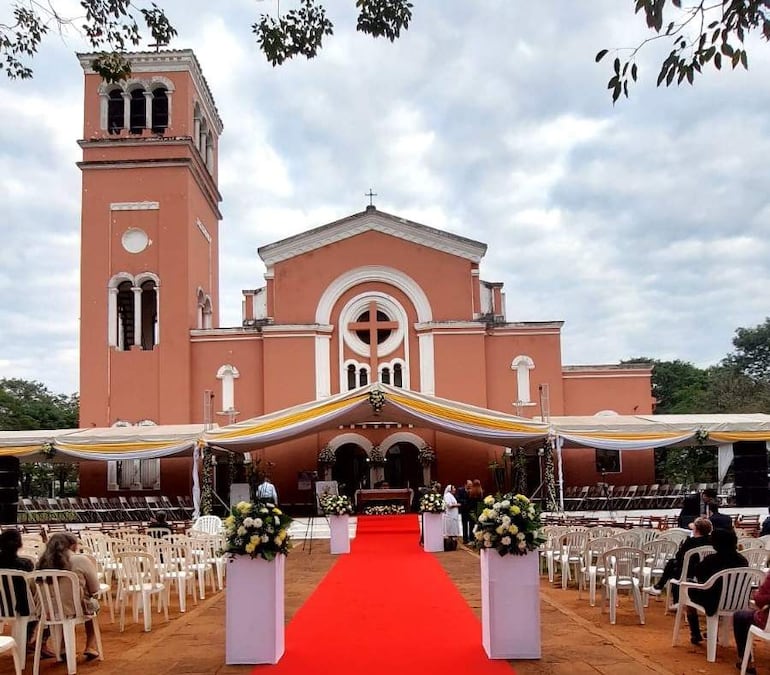 Fachada de iglesia rosada con alfombra roja, sillas blancas y arreglos florales, en ambiente nublado.