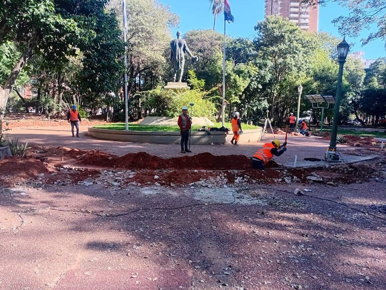 Varios trabajadores con chalecos naranjas y cascos trabajan en un parque con árboles y una estatua al fondo.