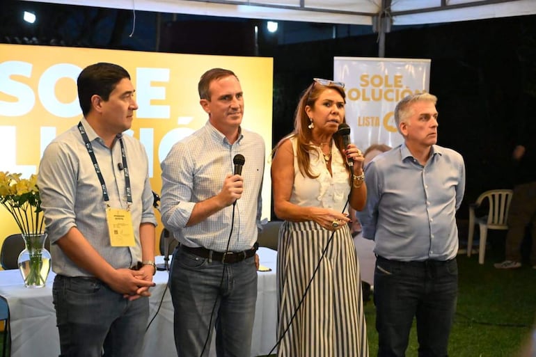 Soledad Núñez con blusa blanca y falda de rayas, sostiene un micrófono mientras tres hombres la observan atentamente en un evento al aire libre.