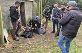 Seis individuos en vestimenta táctica rodean a un hombre inmovilizado, mientras uno sostiene un arma. Fondo de vegetación y pared de ladrillo.